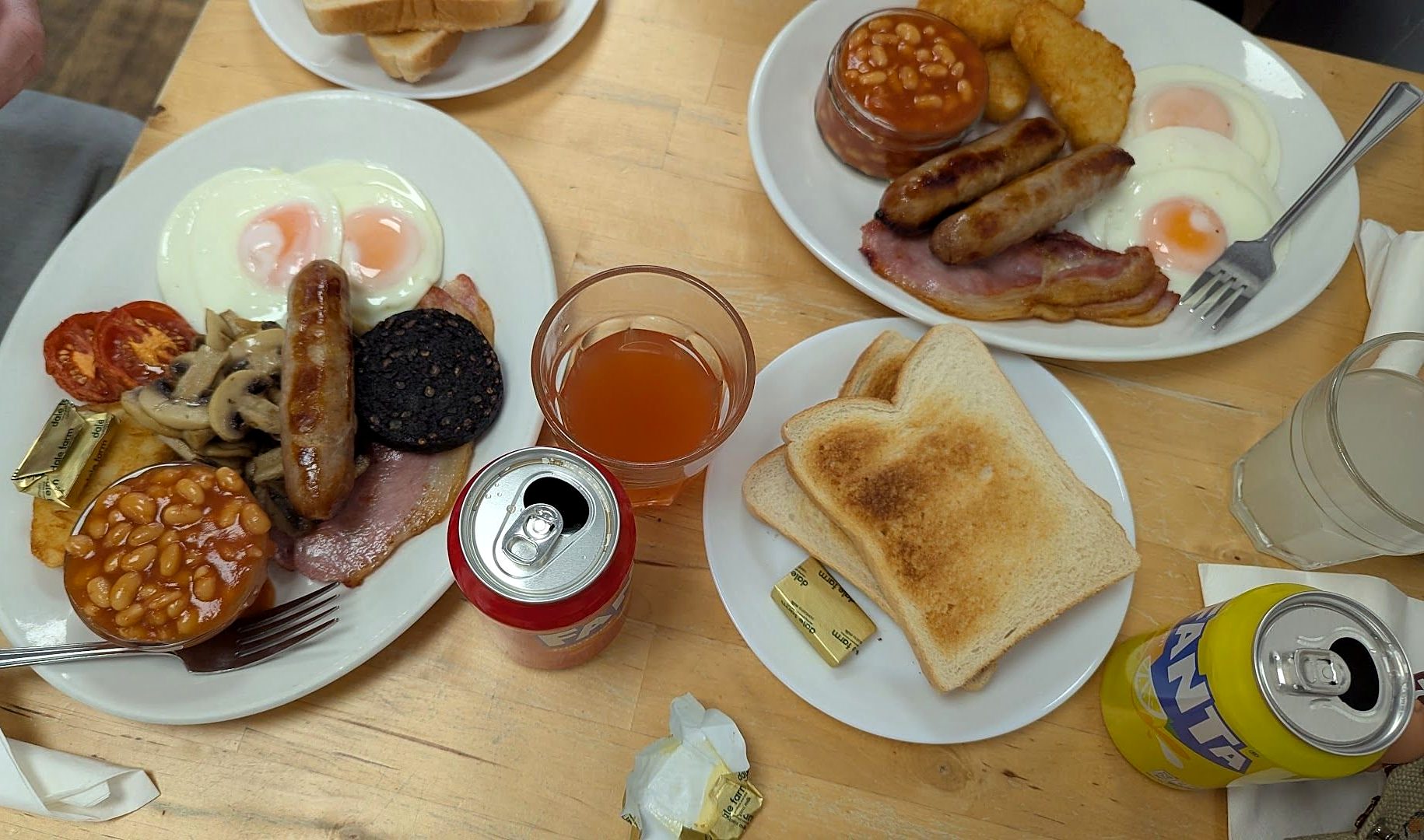 Two full English breakfasts on a wooden table — eggs, sausages, bacon, black pudding, hash browns, mushrooms, tomatoes and beans — with a plate of toast